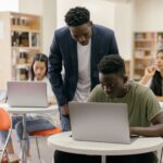 Students learning in a classroom setting with a teacher assisting and laptops on desks, creating an interactive educatio