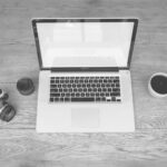 Monochrome image of a laptop, camera, lens, and coffee cup on a wooden desk
